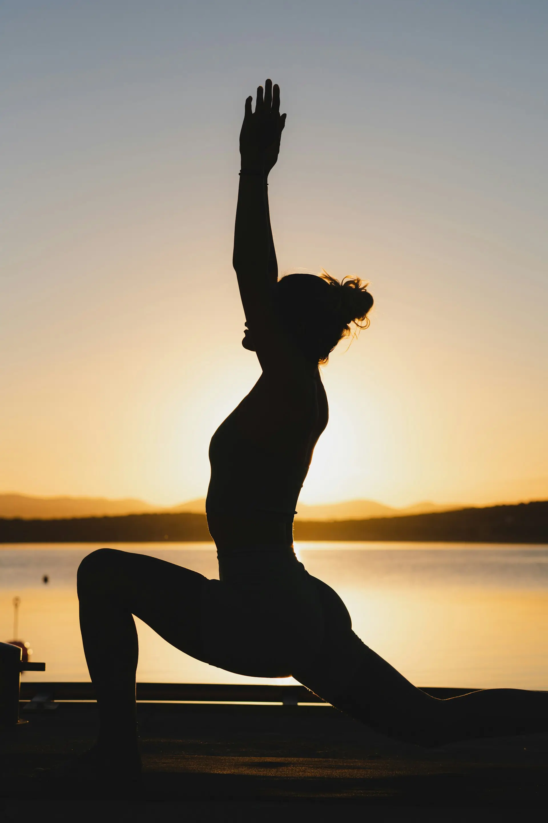 A woman in crescent pose, silhouetted against the sunrise over still water.