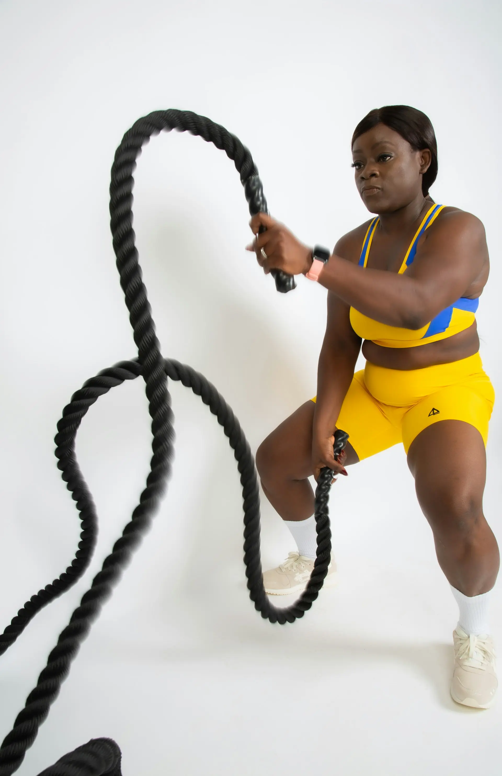 A woman in bold yellow driving battle ropes in a white studio.