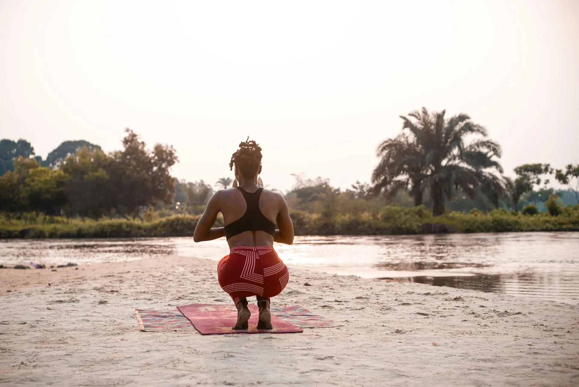 A woman in a deep yogi squat on a mat by the river, palms together.